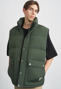 Roheline paks vest kõrge kraega, nööbiklõpsud ja kaks esikotiga. Omab sileda, tikitud tekstuuri ja logomärki.