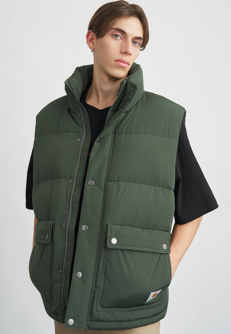 Roheline paks vest kõrge kraega, nööbiklõpsud ja kaks esikotiga. Omab sileda, tikitud tekstuuri ja logomärki.