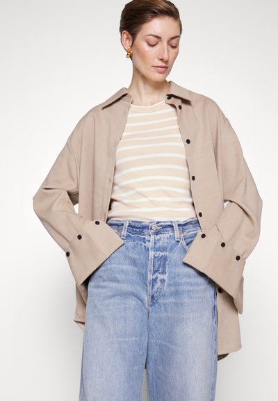 Chemise beige oversize avec des boutons noirs, superposée sur un top crème à rayures. Jean taille haute en denim clair avec une coupe décontractée.