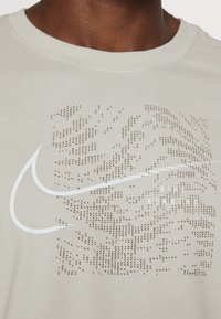 Jasnobeżowa koszulka Nike z okrągłym, teksturowanym wzorem, białym logo oraz gładkim, okrągłym dekoltem. Materiał bawełniany.