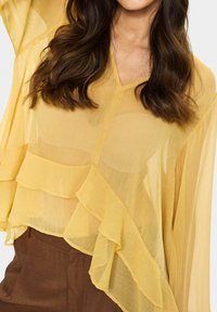 Sheer yellow blouse with a V-neck and layered ruffle design along the hem. Lightweight fabric with a smooth texture. Paired with brown bottoms.