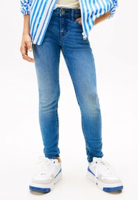 Vaqueros ajustados de color azul con un lavado medio, que presentan una cintura de altura media y un desgaste mínimo. Combinados con zapatillas blancas con acentos azules.
