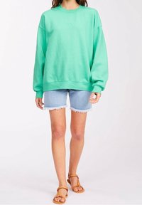Person som bär en lös grön sweatshirt, ljusblå slitna denimshorts och bruna platta sandaler, står mot en vit bakgrund.