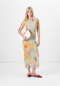 Vrouw in een kleurrijke bloemen midi-jurk, met een witte clutch en rode puntschoen-sandalen, staande tegen een witte achtergrond.