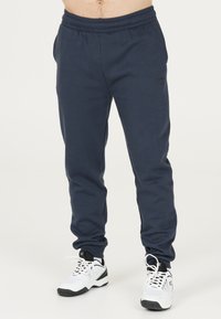 Marina blå sweatpants med elastiskt midjeband, sidofickor och en diskret logodetalj framtill, tillsammans med vita sneakers.