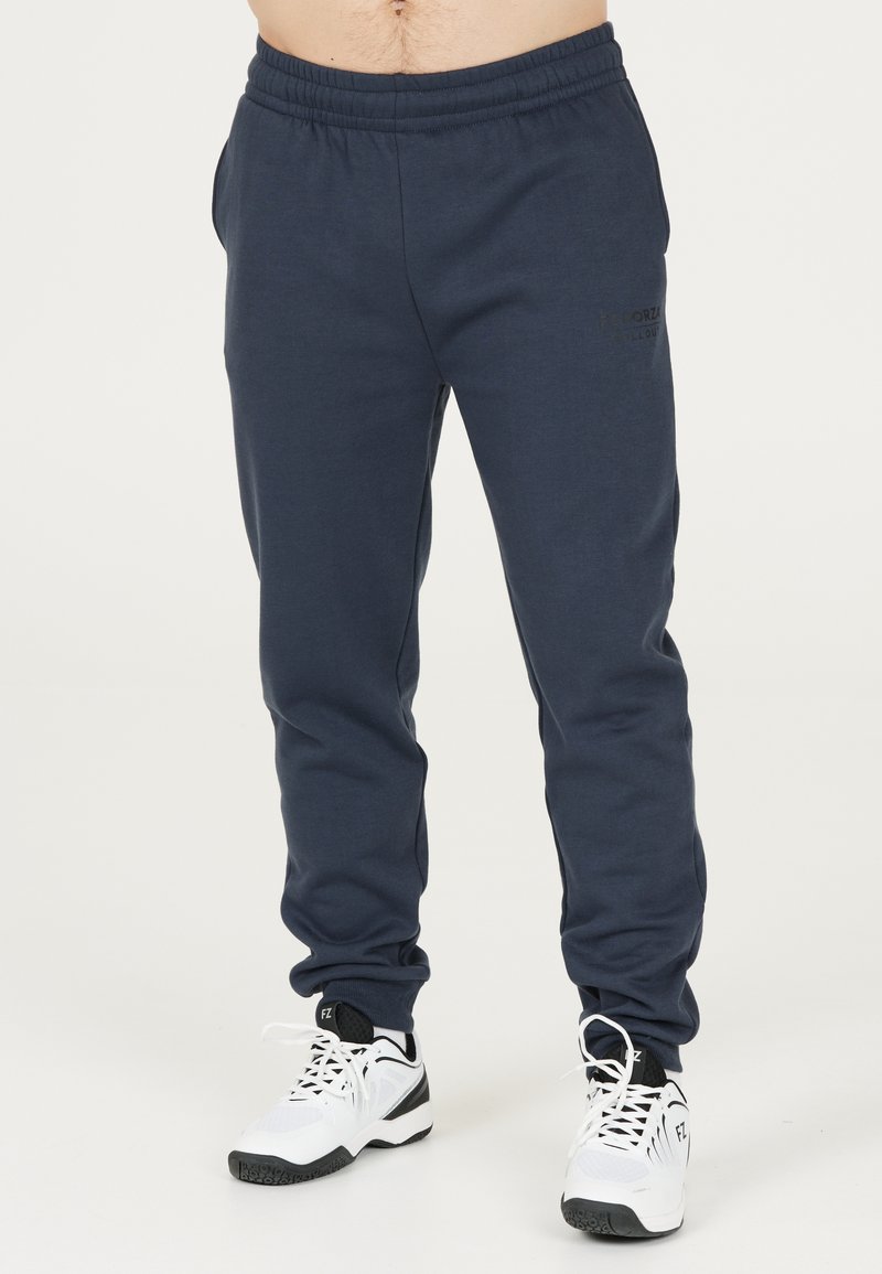 Marina blå sweatpants med elastiskt midjeband, sidofickor och en diskret logodetalj framtill, tillsammans med vita sneakers.