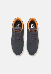 Dark gray suede sneakers with orange accents, round toe, flat rubber sole, and six eyelet lace-up design. Branding visible on the tongue.