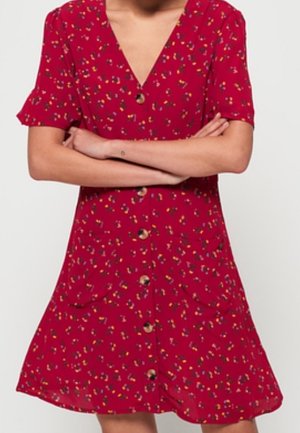 Femme portant une robe rouge fleurie boutonnée avec manches courtes et les bras croisés devant un fond uni.