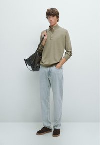 Lys beige sweater med en høj krave og tre knapper, parret med lyseblå jeans og mørkebrune sko. En sort taske hænger over skulderen.