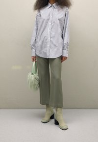 Donna che indossa una camicia a righe blu e bianca con bottoni, pantaloni cropped verde salvia, stivaletti a calza abbinati con tacco e tiene in mano una soffice borsa verde menta.