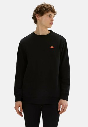 Junger Mann mit lockigem Haar, der ein schwarzes Sweatshirt mit langen Ärmeln und einem kleinen roten und orangefarbenen Logo auf der Brust trägt, schaut seitlich.