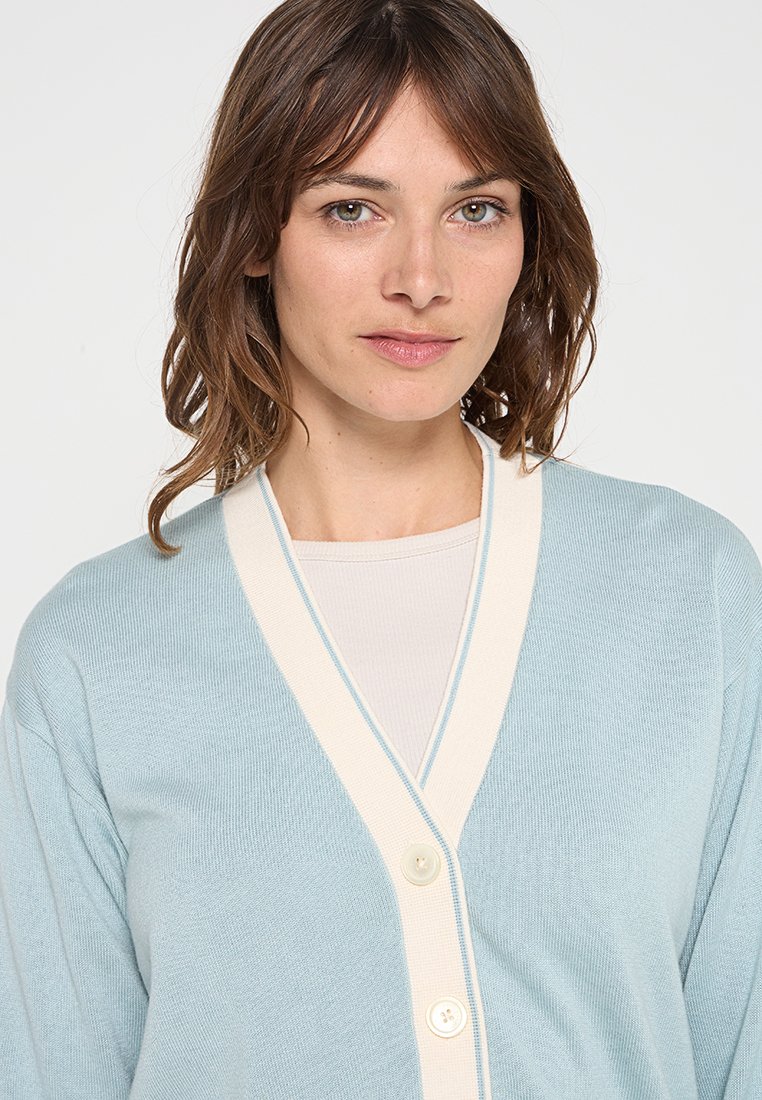 Cardigan azzurro chiaro con scollo a V, dettagli color crema e tre bottoni. La trama sembra morbida e lavorata a maglia, progettata per il comfort.