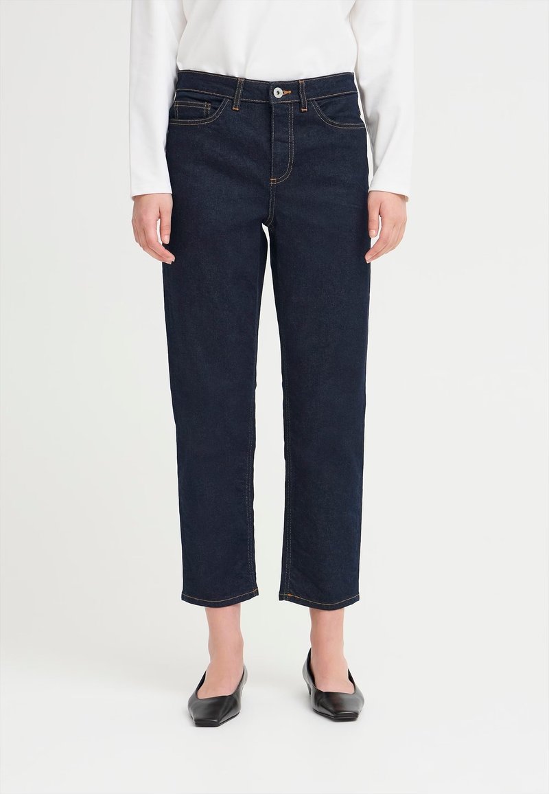 Persona vestida con jeans rectos de color azul oscuro, una blusa blanca de mangas largas y zapatos planos negros de punta, sobre un fondo blanco liso.