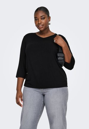 Zelfverzekerde vrouw die een zwart 3/4 mouw top draagt, lichtgrijze spijkerbroek, zilveren oorbellen, en een kleine zwarte schoudertas vast houdt.