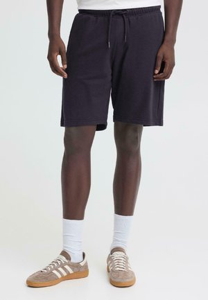 Mężczyzna ubrany w czarne szorty ściągane sznurkiem, białe skarpety za kostkę i jasno-brązowe sneakersy z białymi paskami, stojący na jednolitym tle.