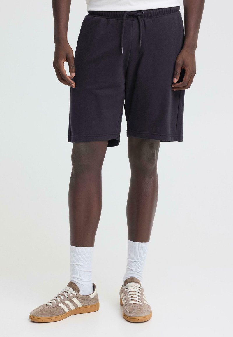 Homme portant un short noir à cordon de serrage, des chaussettes blanches montantes et des baskets marron clair avec des rayures blanches, debout sur un fond uni.