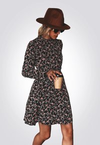 Zwart bloemenjurk met lange mouwen, voorzien van roze bloemen, een hoge nek en een getailleerde taille. Een bruine hoed en een zonnebril complementeren de outfit.