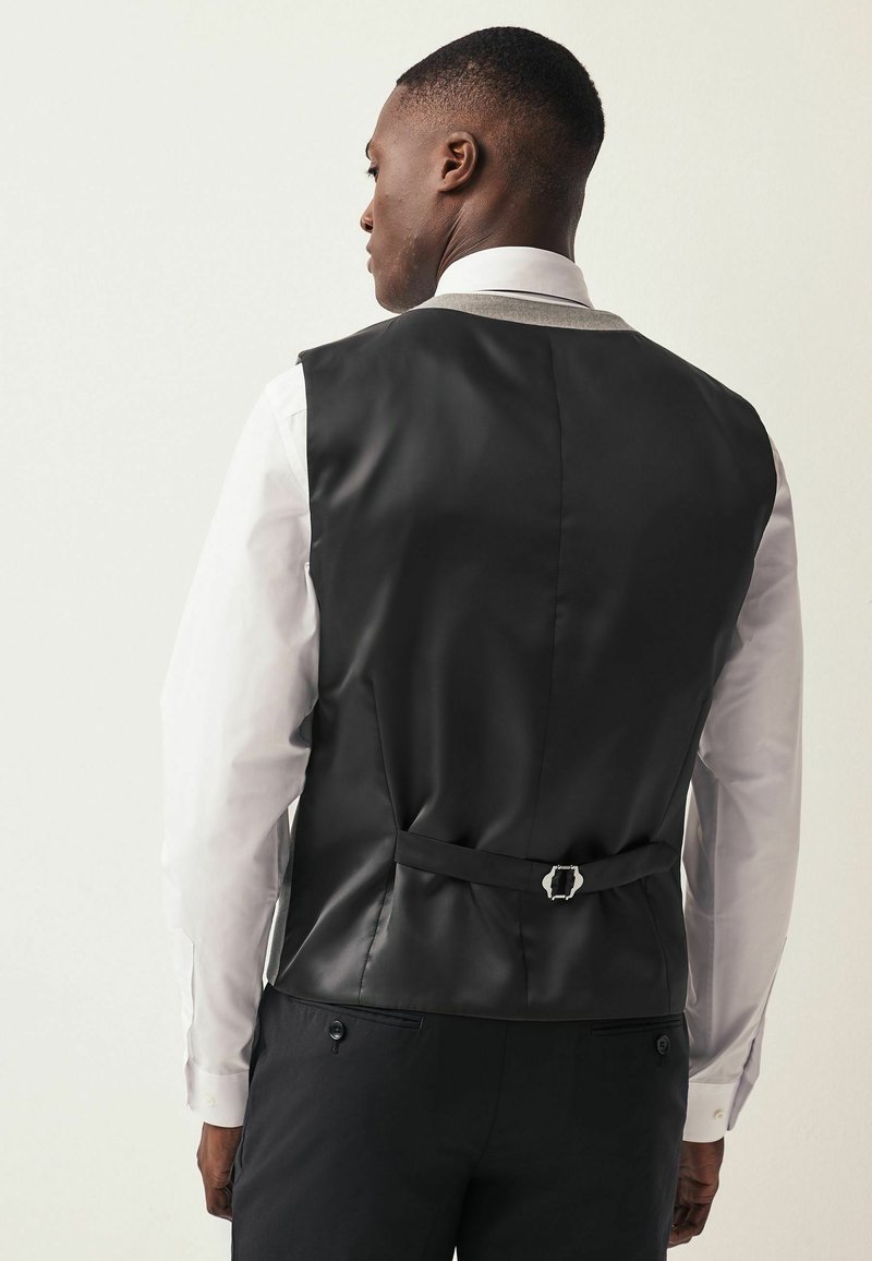Gilet noir et brillant avec un design structuré, présentant un détail de boucle de ceinture au dos et une chemise blanche en dessous.