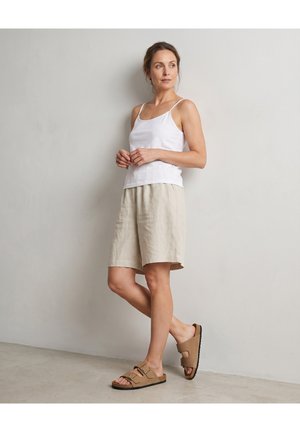 Kvinde står op ad en væg iført en hvid ærmeløs top, beige knælange shorts og brune sandaler med spænde.