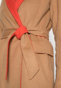 Abrigo de lana beige con un interior de cuello y cinturón rojos. La tela tiene una textura suave con costuras visibles a lo largo de los bordes.
