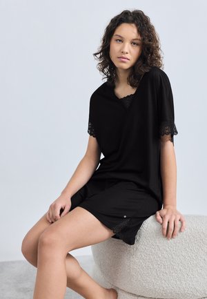 Ung kvinde med krøllet hår iført en sort top med blondekanter og shorts, siddende på en struktureret hvid skammel mod en ensfarvet baggrund.