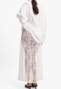 Falda maxi blanca de encaje con patrón floral, superpuesta sobre una base sólida. Combinada con una camisa blanca holgada y sandalias de tacón.