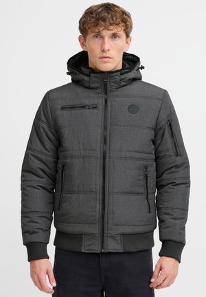 Veste matelassée en tissu gris foncé avec une finition texturée. Comprend une capuche amovible, de multiples poches zippées et des poignets côtelés.