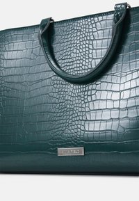 Groene handtas met krokodillenmotief, gemaakt van glanzend synthetisch materiaal. Voorzien van stevige handvatten en een metalen merkembleem.