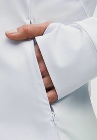 Light gray athletic jacket with a pocket on the side, featuring a concealed zipper and a snug cuff that partially covers a hand.