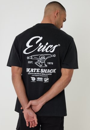 Homme vu de dos, les mains derrière le dos, portant un t-shirt noir imprimé avec "Erics Skate Shack" et un graphique de truck de skateboard en blanc.