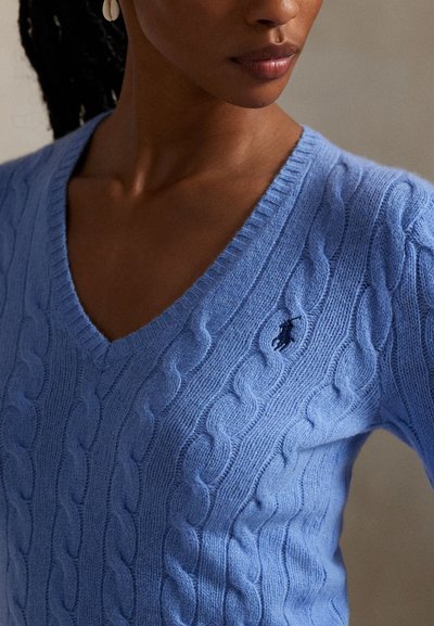 Nő világoskék fonott mintás V-nyakú pulóverben, amelynek mellrészén kis hímzett logó található, és kagylófülbevalót visel.