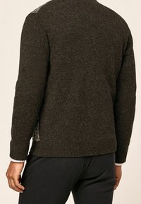 Uomo che indossa un maglione di maglia marrone scuro e pantaloni neri, mostrato di spalle con le mani rilassate ai lati.