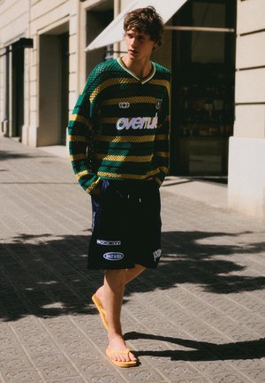 Ung mand går på byens gade iført en grøn og gulstripet sweater, sorte shorts med logoer og gule flip-flops.