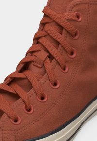 Κοντινή λήψη μονοπατιού σουέντ σε σκουριασμένο χρώμα, με χονδρά κορδόνια στο ίδιο χρώμα περασμένα από επτά οπές στη μία πλευρά.