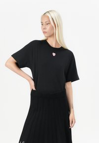 Maglietta nera a maniche corte con scollatura rotonda e una piccola toppa a forma di cuore. Indossata con una gonna nera plissettata, caratterizzata da un elastico a coste in vita.