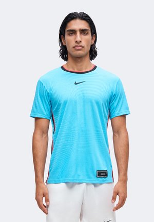 Homme aux cheveux noirs mi-longs portant un maillot de sport Nike bleu clair à manches courtes et un short blanc, debout devant un fond uni.