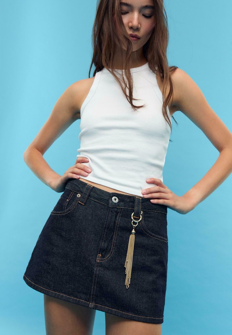 Jupe en denim bleu foncé avec coutures visibles, deux poches avant et un accent de gland doré sur la boucle de ceinture. Haut sans manches blanc.