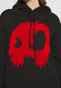 Sweat à capuche noir avec un grand graphique rouge ressemblant à un visage stylisé et poilu de créature sur la poitrine, porté par une personne.