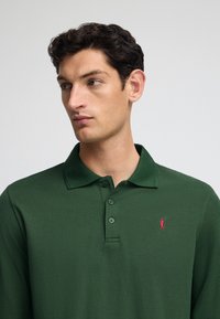 Grünes Polohemd aus strukturiertem Stoff, mit einer Drei-Knopf-Leiste und einem kleinen roten Logo auf der linken Brust.