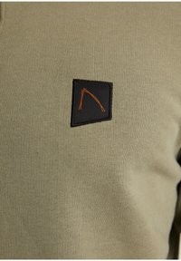 Khaki košile s dlouhým rukávem a elastickou strukturou; obsahuje černý čtvercový nášivku vyšitý s oranžovým designem.