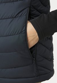 Izolirani puffer prsluk, tamnoplave boje, s patentnim zatvaračem. Tkanina izgleda glatko s detaljima u obliku kvadrata.