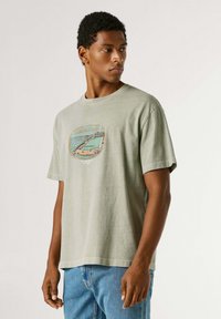 Jonge man in lichtgrijs T-shirt met print en blauwe spijkerbroek, kijkt opzij tegen een effen achtergrond.