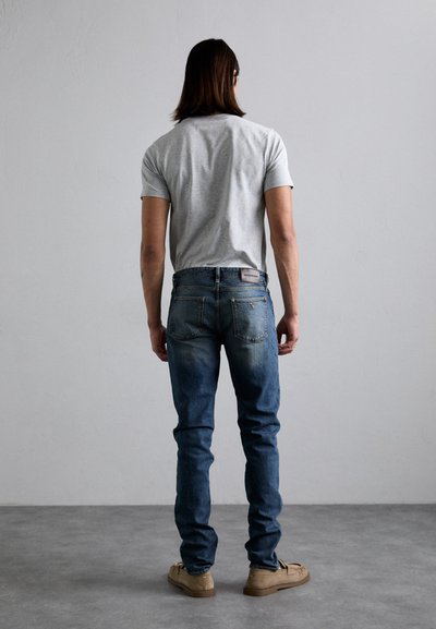 Camiseta cinza de mangas curtas combinada com jeans de denim azul, com um leve desbotado, bolsos traseiros e uma etiqueta de couro marca. Sapatos castanhos.