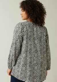 Donna con capelli ricci che indossa una blusa a maniche lunghe bianca e nera a pois, girata leggermente di lato su uno sfondo semplice.