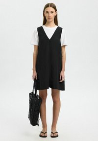 Zwarte linnen jurk met een diepe V-hals, gelaagd over een wit t-shirt. Gecombineerd met een zwarte mesh schoudertas en zwarte slippers. Eenvoudig ontwerp.