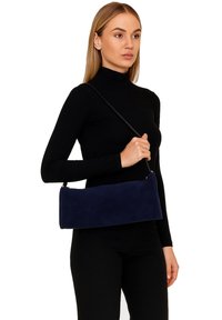 Navyblauwe suède clutch met een rechthoekige vorm, korte schouderband en strakke lijnen, vastgehouden door een persoon die een zwarte coltrui draagt.