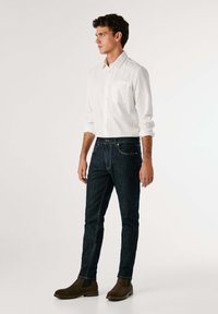 Bijela košulja s dugim rukavima i džepom na prsima, uparena s tamnoplavim trapericama i smešnim suede Chelsea čizmama. Slim-fit silueta, osnovni dizajn.