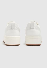 Un paio di sneaker bianche basse in pelle testurizzata, con suole spesse di un bianco sporco e linguette sul tallone, viste dal retro su uno sfondo neutro.