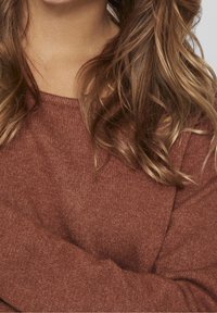 Maglione di colore ruggine con scollatura a girocollo, tessuto testurizzato e maniche lunghe, mostrato parzialmente con le braccia incrociate.