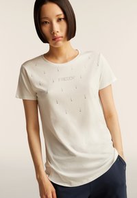 Camiseta blanca de manga corta con la palabra "FREDDY" en tachuelas plateadas, con una textura suave y un corte relajado. Detalles de tachuelas a lo largo.